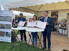 Deux hommes et une femme se tiennent derrière deux tables hautes et brandissent, rayonnants, trois exemplaires d'un accord. On aperçoit le stand du FiBL à l'arrière-plan.