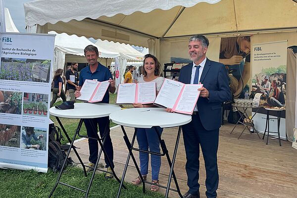 Two men and a woman stand behind two bar tables, beaming and holding up three copies of an agreement. The FiBL stand can be seen in the background.