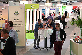 The picture shows a corridor with promotional stands on either side. There are several bar tables around which people are standing and chatting. Everyone is wearing a green exhibition badge with a name tag around their neck. A waiter can also be seen among the crowd.