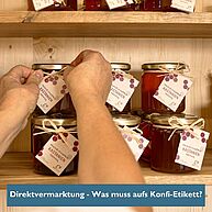 Zwei Hände binden an einem Maroni-Konfitürenglas ein Etikett an, das Glas steht mit weiteren Gläsern in einem Regal.