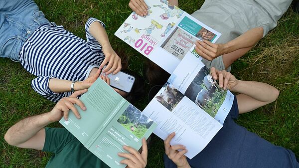 Four people lie on the grass holding a magazine, book, leaflet and smartphone.