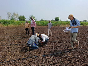Several men work together in a cultivated field; two kneel on the ground while another stands next to them with notes.
