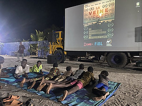 Personen liegen im Freien auf Decken im Sand und schauen einen Film, die Leinwand ist an einem LKW angebracht.