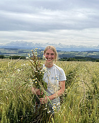 Eine Frau steht in einem Weizenfeld und hält einen Beikraut-Strauss in die Kamera.
