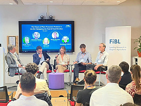 The seven participants of the roundtable discussion sit in a semi-circle in front of an audience. The participants' professional profiles are displayed on a blue screen in the background.