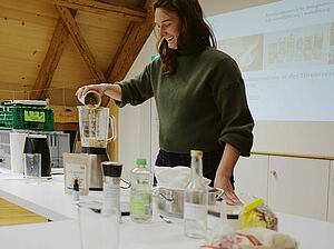 Frau an Tisch, die Zutaten in einen Mixer füllt. 