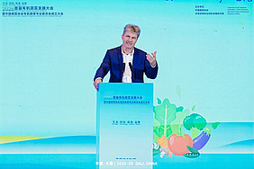 A man is standing behind a lectern, speaking. The background is a bright turquoise colour.