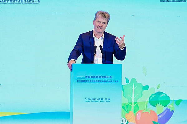 A man is standing behind a lectern, speaking. The background is a bright turquoise colour.