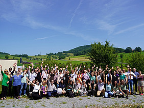 A group of around 50 people on a sunny roof with green hills in the background.