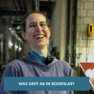 Eine junge Frau lacht aus vollem Herzen. Sie trägt einen einfachen Pullover, welcher Erdflecken von der Arbeit drauf hat. Sie hat eine Brille und braune Haare als Pferdeschwanz. Im Hintergrund ist ein Labor zu sehen. Banner "Was geht ab im Bodenlab?"