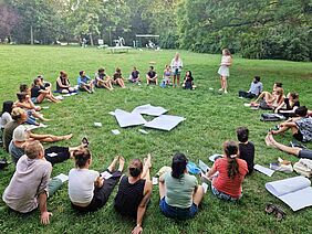 About 30 people sitting in a circle on a meadow. 