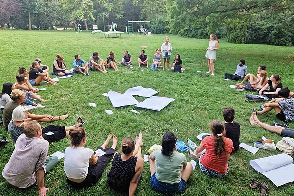 About 30 people sitting in a circle on a meadow. 