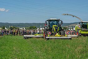 Traktor mit Mähwerk, daneben Mähdrescher, im Hintergrund grosse Menschengruppe