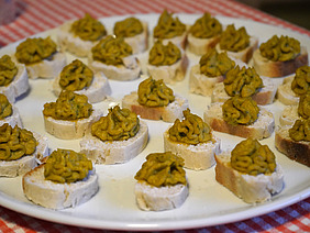 Auf einem Teller sind in Kreisen viele kleine Brotscheiben angeordnet. Jede Brotscheibe hat einen grosszügigen, schönen Spritzer gelbgrünen Hummus darauf.