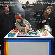 Eine Person sitzt an einem Tisch vor bunten Bausteinen. Sie ist um die 50, trägt eine bunt-gestreifte Mütze und schaut verspielt und kritisch auf die Bausteine. Zwei junge Leute stehen daneben und schauen neugierig und belustigt zu. Im Hintergrund ist eine vollgeschriebene Wandtafel. Die Stimmung ist kreativ und angeregt.