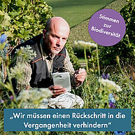 Ein Mann um die 50 hockt in einer Blumenwiese mit einem Tablet in der Hand und ist in die Beobachtung von Etwas vertieft. Er hat eine Glatze, trägt ein kariertes Hemd und eine Weste darüber und schaut sehr konzentriert.