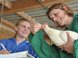 One woman examines the hen's beak. The other takes notes.