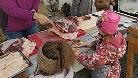 Zwei Kinder an einem Tisch, eine Person zeigt auf ein Stück Fleisch