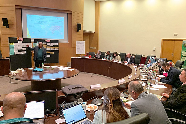 Une personne debout devant un tableau d'affichage, face à un demi-cercle de personnes assises.