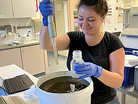 Junge Frau mit Pipette und Glasgefäss in den Händen steht hinter einem Eimer gefüllt mit brauner Flüssigkeit.