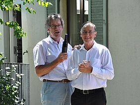 Deux hommes sourient à la caméra, l'homme de gauche tient une bouteille de vin.