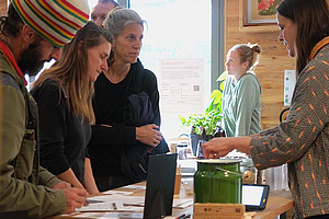 Drei Leute und noch mehr im Hintergrund stehen an einem Stand am Bioleguminosentag. Der vordere Mann trägt eine auffällige, grosse, gestrickte Mütze. Eine Frau steht hinter dem Stand. Sie sprechen miteinander und die Gäste schauen neugierig.