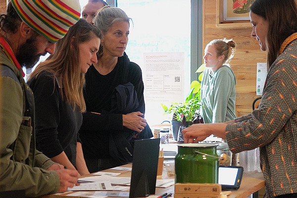 Drei Leute und noch mehr im Hintergrund stehen an einem Stand am Bioleguminosentag. Der vordere Mann trägt eine auffällige, grosse, gestrickte Mütze. Eine Frau steht hinter dem Stand. Sie sprechen miteinander und die Gäste schauen neugierig.