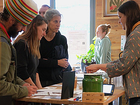 Drei Leute und noch mehr im Hintergrund stehen an einem Stand am Bioleguminosentag. Der vordere Mann trägt eine auffällige, grosse, gestrickte Mütze. Eine Frau steht hinter dem Stand. Sie sprechen miteinander und die Gäste schauen neugierig.