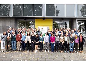 A group of roughly 50 people standing in front of a building, smiling.