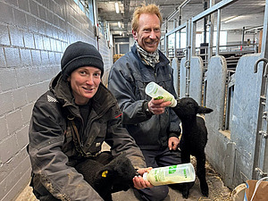 Zu sehen sind eine Frau und ein Mann in Arbeitskleidung, die jeweils ein Lamm mit einer Flasche füttern.