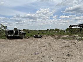 Im Hintergrund eine Wiese mit Kühen, im Vordergrund ein zerstörter Panzer und das Gerüst eines Hauses.