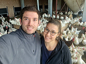 Zwei junge Leute um die dreissig schauen lächelnd in die Kamera. Sie umarmen sich und sind offensichtlich ein Pärchen. Im Hintergrund sind viele Hühner in einem Stall zu sehen.