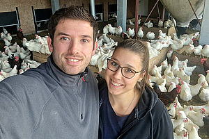 Zwei junge Leute um die dreissig schauen lächelnd in die Kamera. Sie umarmen sich und sind offensichtlich ein Pärchen. Im Hintergrund sind viele Hühner in einem Stall zu sehen.