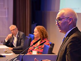 Close up of a speaker, behind him a woman and a gentleman at a longer lectern