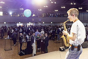Saxophon player on stage, people at standing desks in the background.
