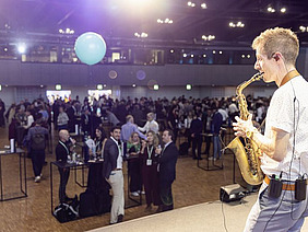 Saxophon player on stage, people at standing desks in the background.