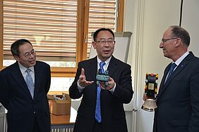 Three men standing, two of them are exchanging gifts (a small Chinese pot and a Swiss cow bell).