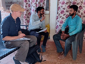 Three men, including a foreign researcher, sit on chairs in a house with a dirt floor and a fan. 