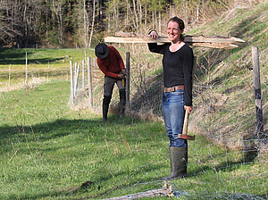 Zu sehen ist eine Frau, die einen Hammer in der Hand hält und Holzpfähle auf der Schulter trägt. Im Hintergrund ist ein Mann zu sehen, der einen Weidezaun errichtet.
