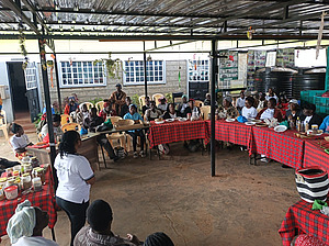 On voit un cercle formé de tables et de chaises sous un auvent. Les chaises sont occupées par des personnes qui écoutent une femme. Cette photo a été prise dans le milieu culturel kenyan.