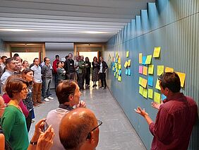 A group of people standing in front of a wall and discussing the various notes that are hung up.