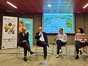 Four people sitting in front of a screen and banners.