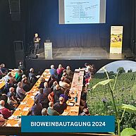 Eine Gruppe von Menschen an Tischen schauen dem Vortrag auf der Bühne zu.