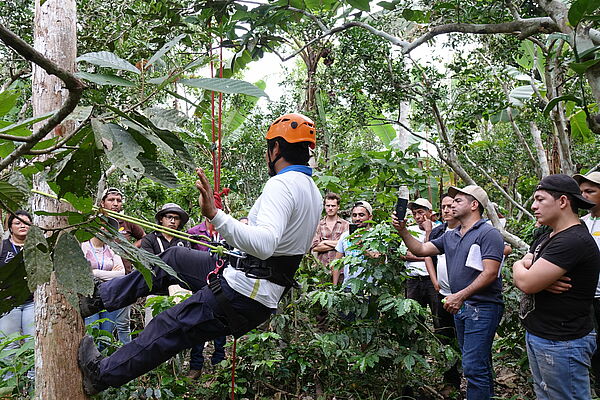 Menschen schauen einem Mann zu, wie er mit Kletterausrüstung einen Baumstamm erklimmt.