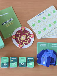 Ein Spiel ist zu sehen: mehrere Karten, ein kleines, rundes Spielbrett, eine Verpackung. Alles in Braun- und Grüntönen.