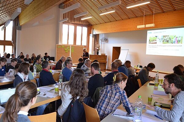 Ein Gruppe von Leuten sitzt in einem Konferenzraum; es läuft eine PowerPoint-Präsentation.