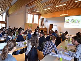 Ein Gruppe von Leuten sitzt in einem Konferenzraum; es läuft eine PowerPoint-Präsentation.