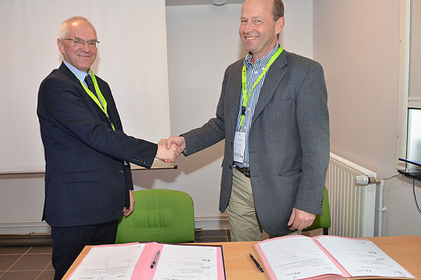 Two men are shaking each other's hands and smiling. In front of them, there are two signed documents on a table.