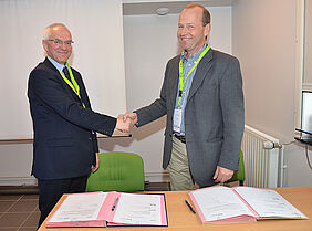 Two men are shaking each other's hands and smiling. In front of them, there are two signed documents on a table.