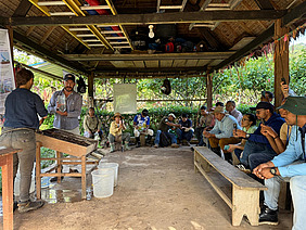 A group of people sitting under a hut. Two people demonstrating an experiment. 